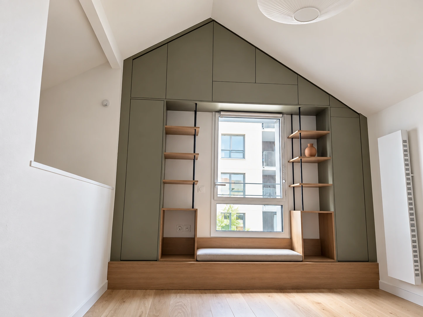 Mur de rangements vert de gris suivant le pignon, étagères bois et banquette autour d’une fenêtre